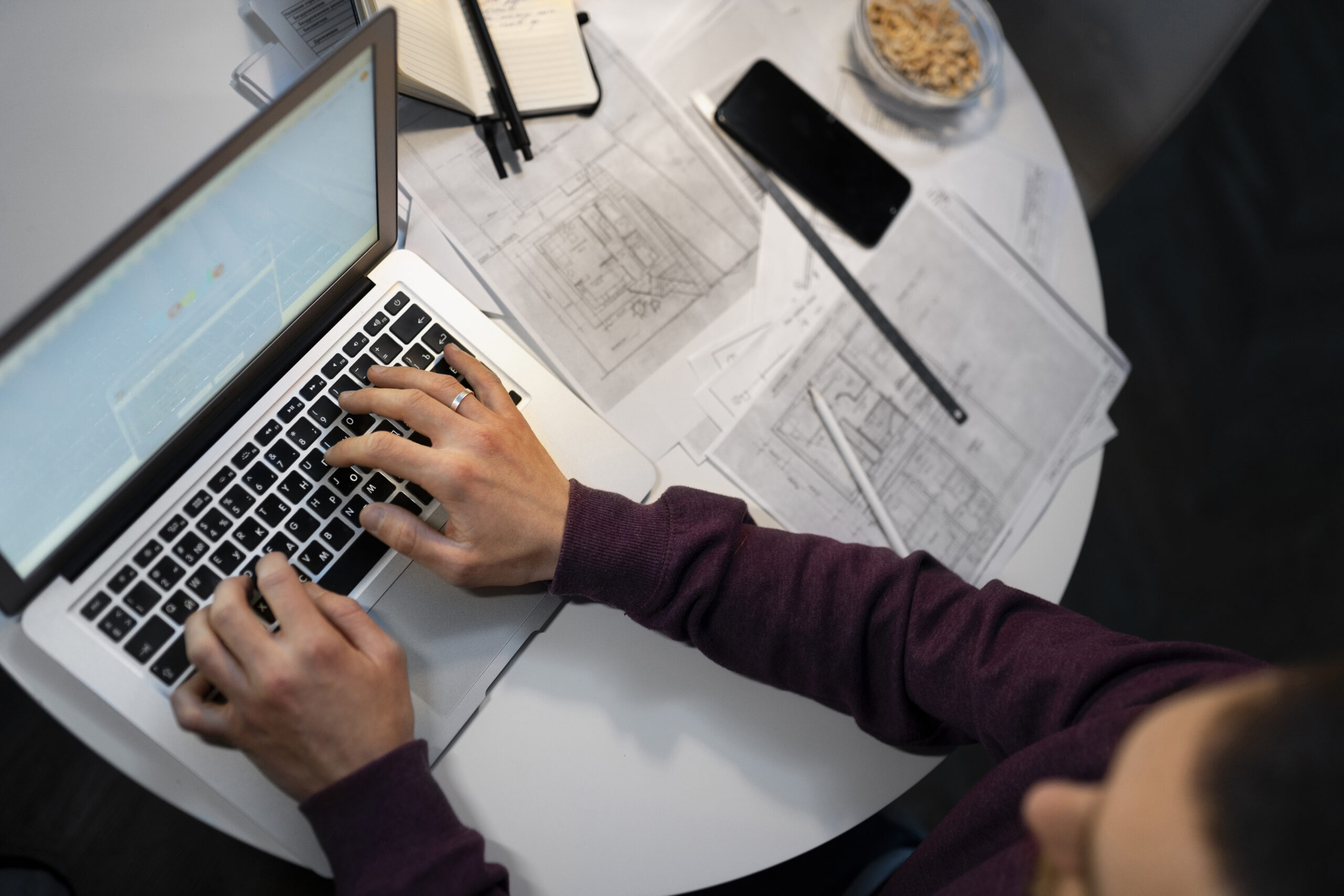 An engineer at a desk works on a laptop surrounded by floor plans, demonstrating troubleshooting common issues in electrical project management software to keep the project on track.