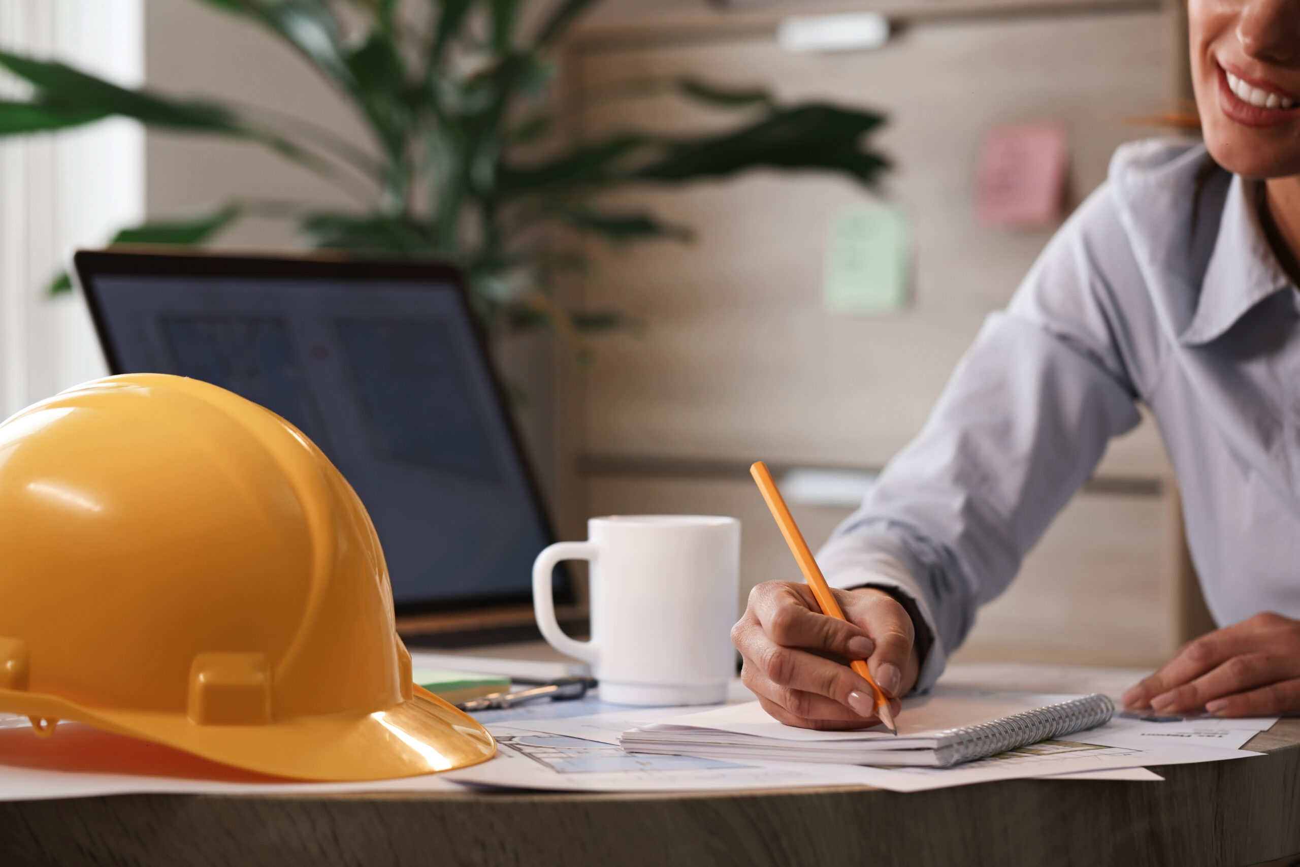 Woman writing notes at a desk with a hard hat and laptop beside her, illustrating an expert checklist for choosing MEP project management software.