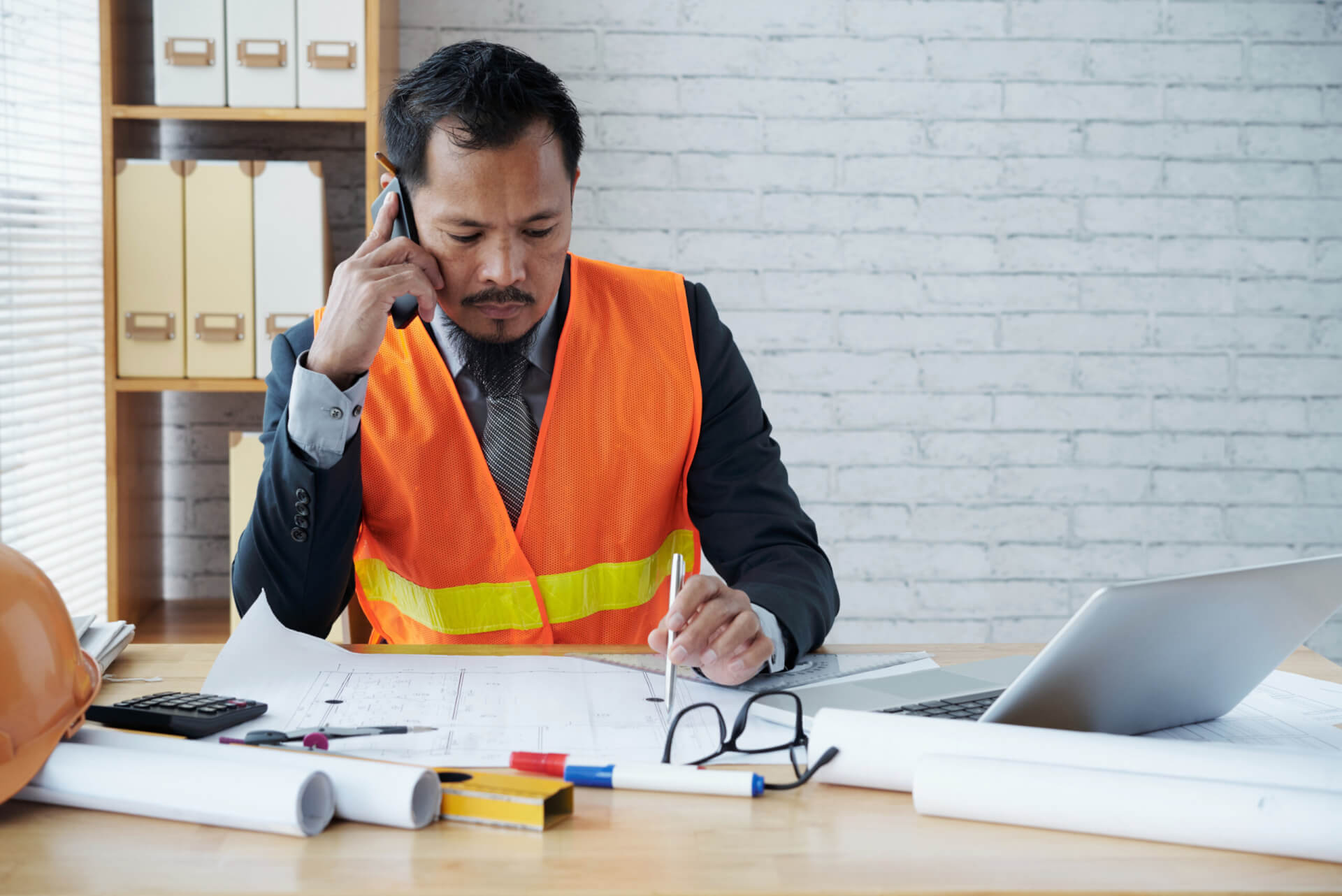 A construction manager on the phone at a desk with blueprints, a laptop and a hard hat, illustrating the process of troubleshooting common issues in mechanical project management software.