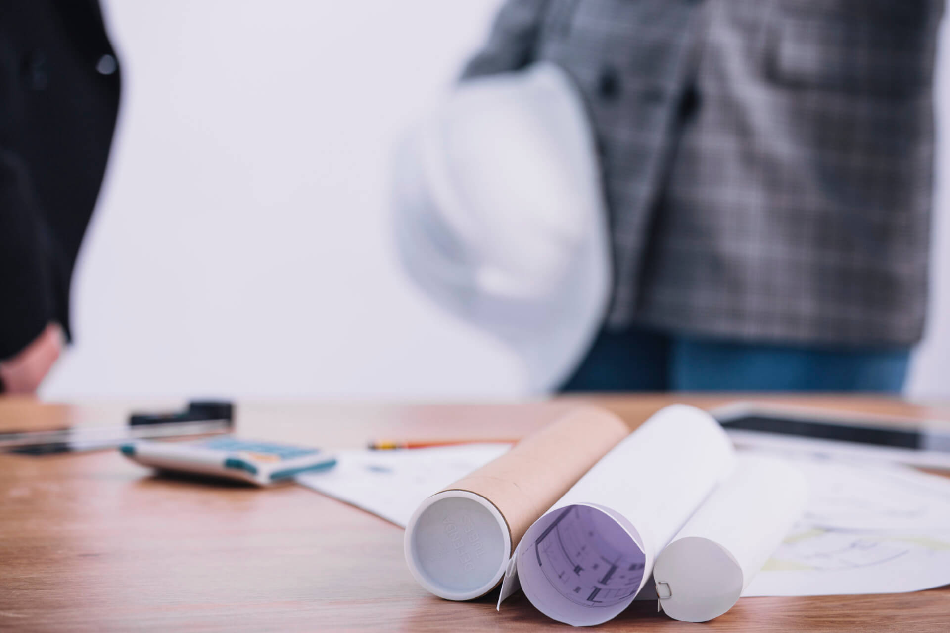 Rolled blueprints and a hard hat on a desk with a contractor in the background, representing time-saving tips for using mechanical project management software.