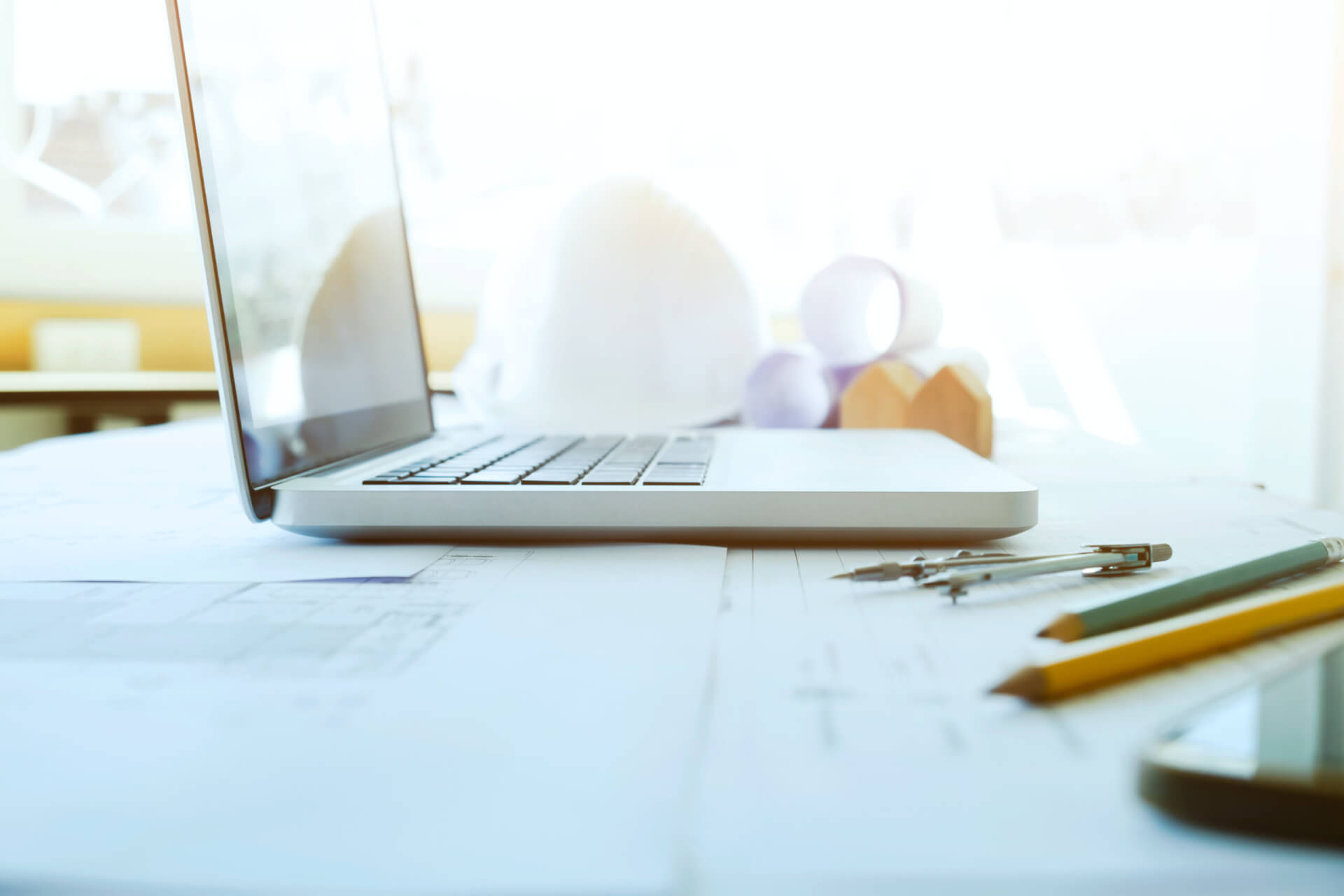 A laptop open on construction drawings with a hard hat and pencils on the desk, representing the pros and cons of cloud-based mechanical project management software.