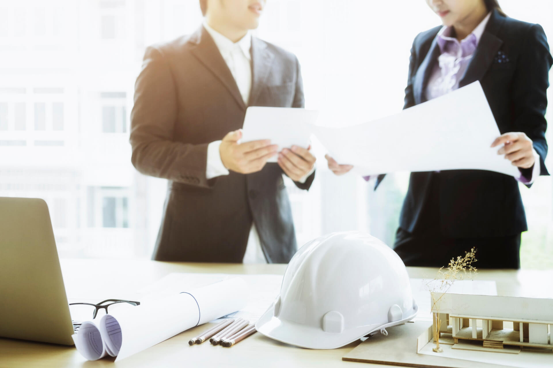 Two engineers reviewing project documents in a meeting with blueprints, a hard hat and a laptop on the desk, illustrating how to implement mechanical project management software across teams.